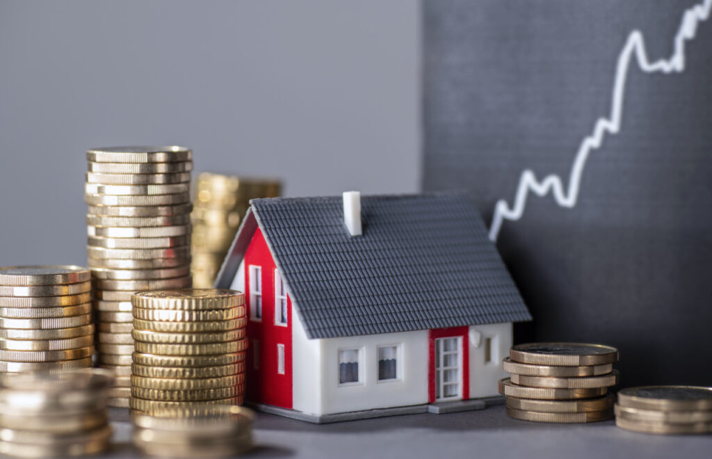  Model house surrounded by stacks of coins with a rising financial chart in the background.